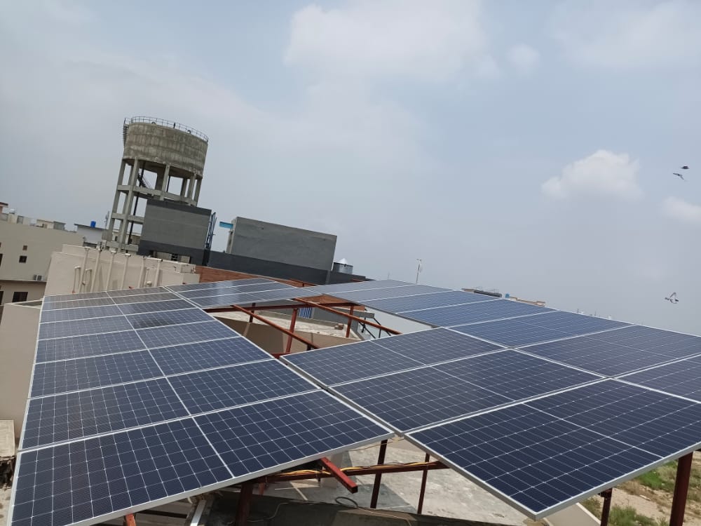 Rooftop solar array on a Pakistani home, mounted on a raised steel structure with a water tank and neighbouring houses behind